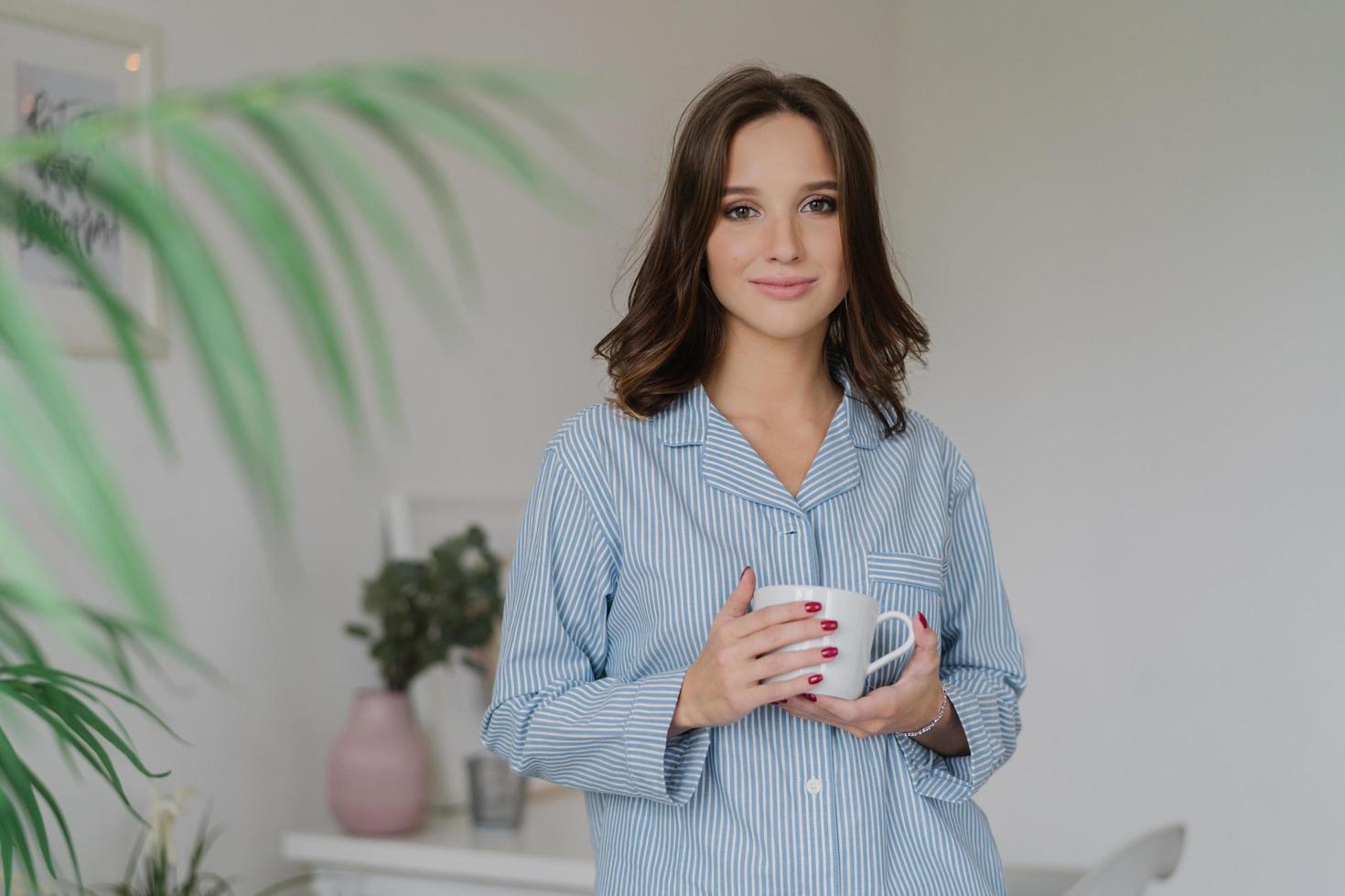 Morning time concept. Young pretty European woman in nightclothes carries white mug of coffee or cappuccino, stands indoor in living room, has weekend, enjoys good rest. People and lifestyle photo