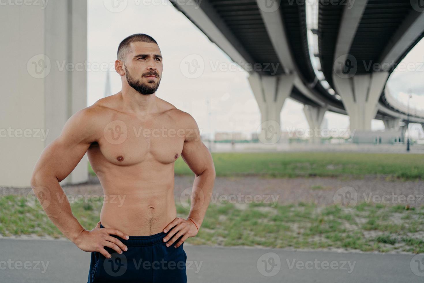 foto de un apuesto hombre barbudo sin camisa con poses de torso desnudo al aire libre mantiene las manos en la cintura concentradas en la distancia va a hacer deporte, hace ejercicio matutino, orgulloso de ser fuerte, tiene abdominales perfectos