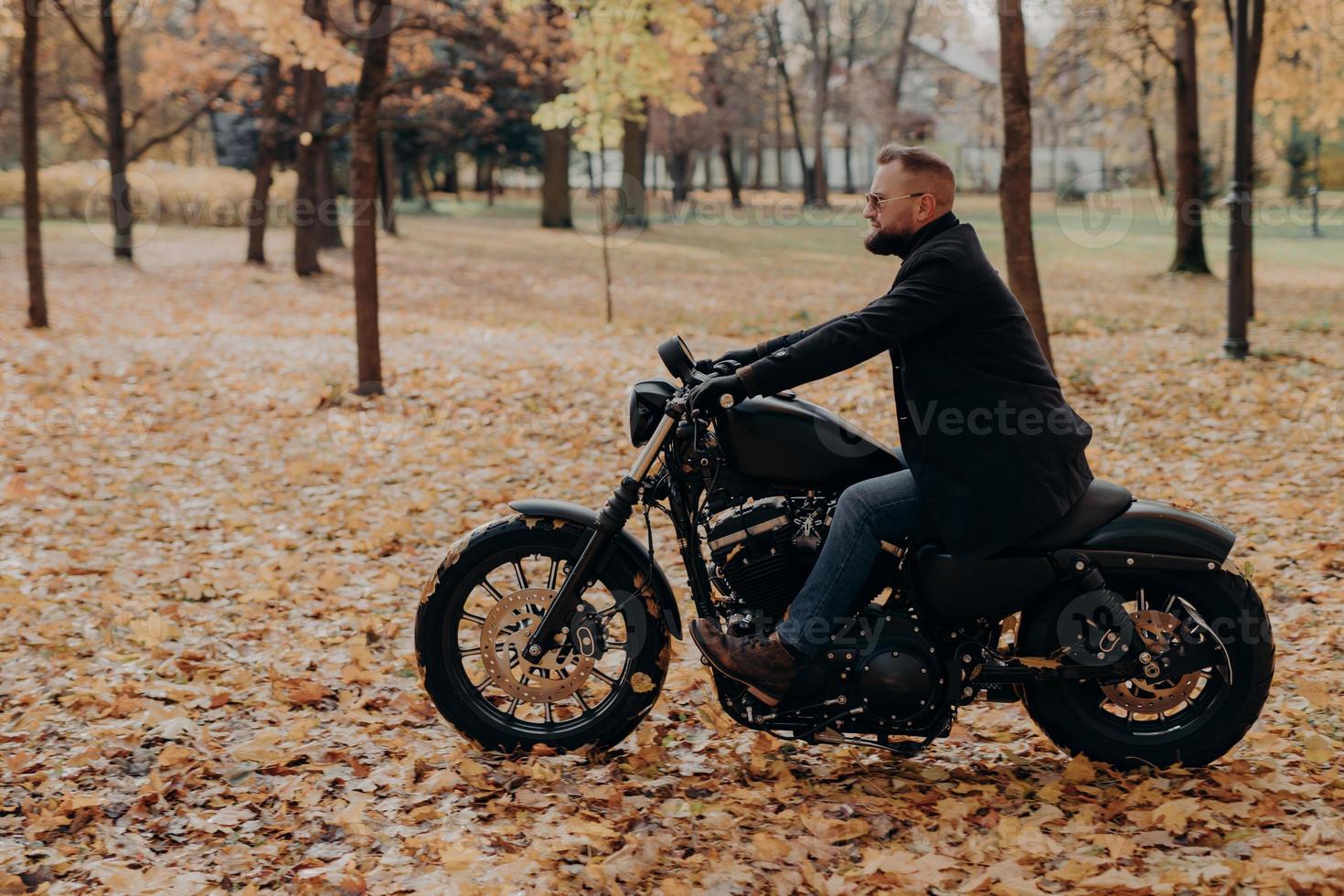 Sideways shot of confident male biker rides motorcycle, has adventure on two wheels in autumn park, wears stylish clothing, protective sunglasses, enjoys nature during beautiful fall season. photo