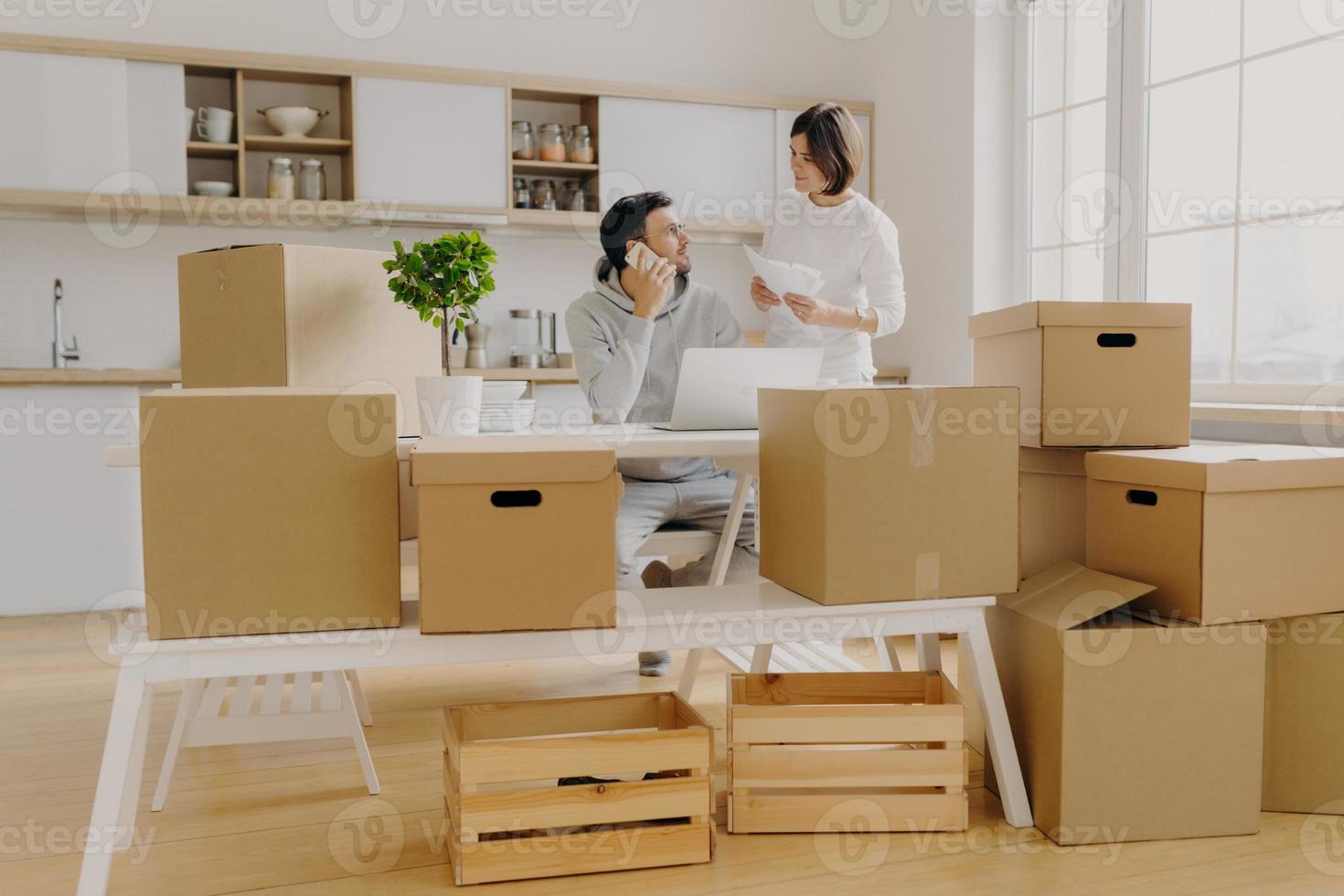 Family review their bank account, get loan application, talk about expenses, man talks via cellphone, sits at desk with laptop computer woman holds papers pose against kitchen interior with big window photo