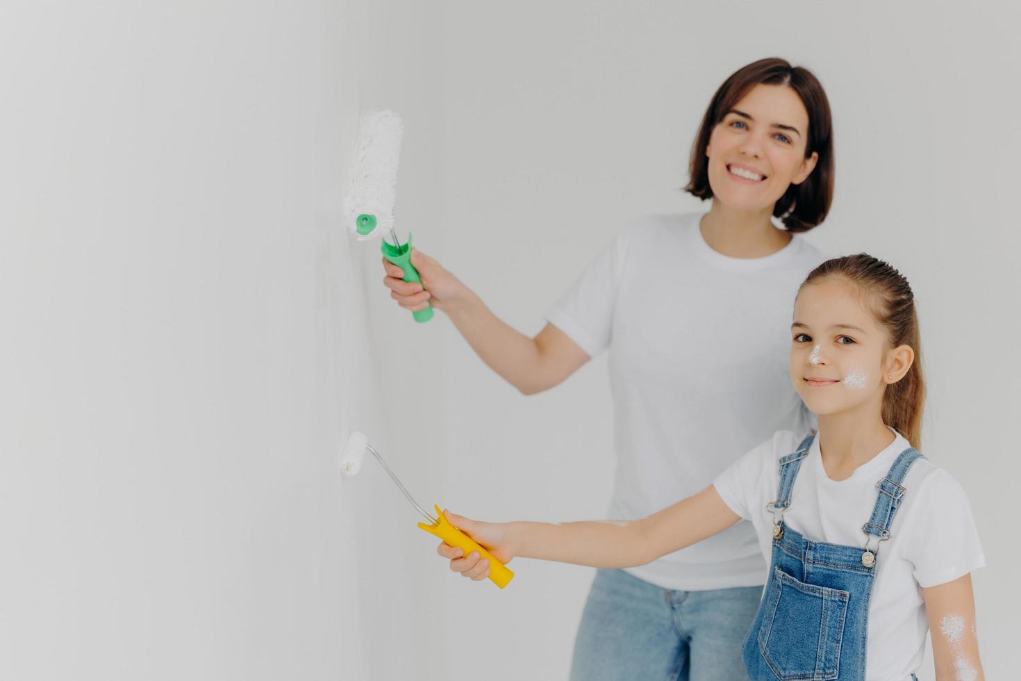 Horizontal shot of happy girl and her mother paint walls in white color, use paint rollers, improve house, busy with domestic work, move in new apartment. Home repair and improvement concept. photo