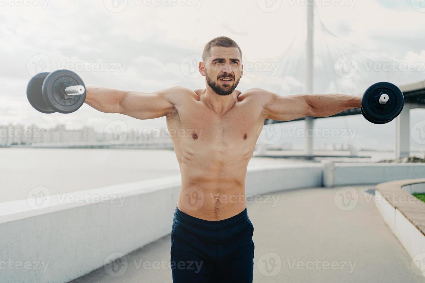 Serious bearded athlete man raises barbells puts all efforts in gaining strong muscles on arms has well built body poses outdoor near river. Determined sportsman has weightlifting exercise outside photo