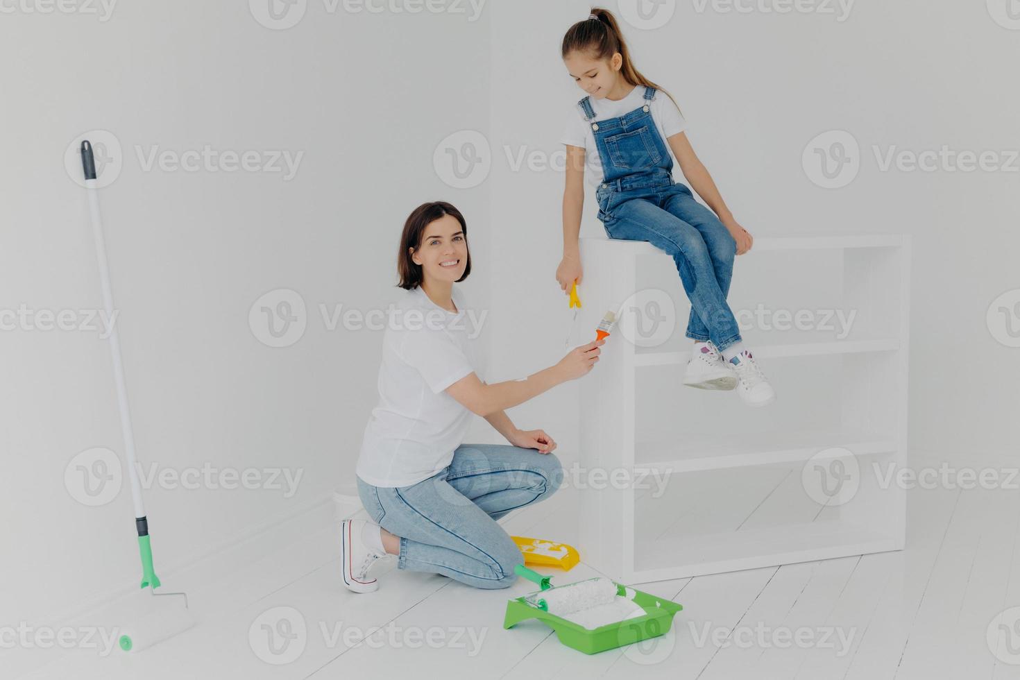 toma horizontal de una madre cariñosa y su hija ocupadas renovando muebles en casa, usan pintura blanca y pincel, rodillo, feliz casi terminando el trabajo, ocupadas pintando una casa nueva, vestidas informalmente foto