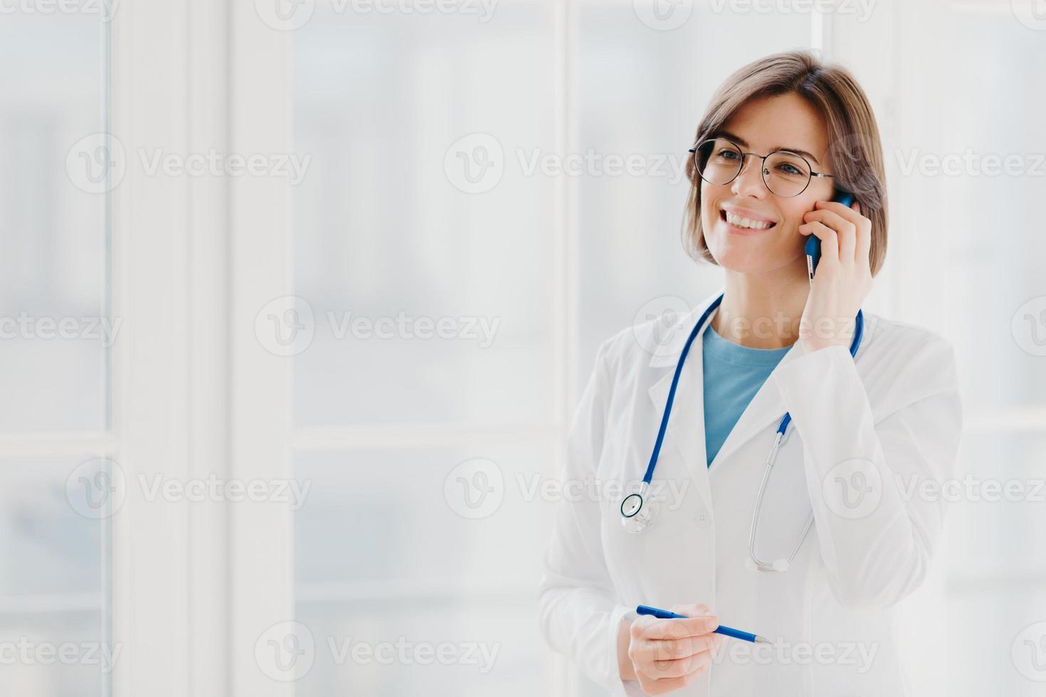 Shot of pleased female physician or surgeon gives prescription via mobile phone, has conversation with patient, holds pen, wears white uniform, works in hospital, stands indoor against window photo