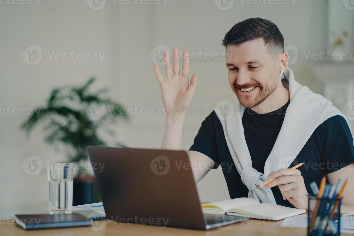 Cheerful bearded man has video conference online waves palm in laptop computer uses wireless earphones poses at coworking space dressed casually writes down information in diary smiles gladfully photo