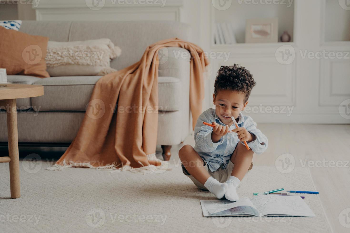 Cute afro american boy kid with colouring book sitting on floor in cozy living room photo