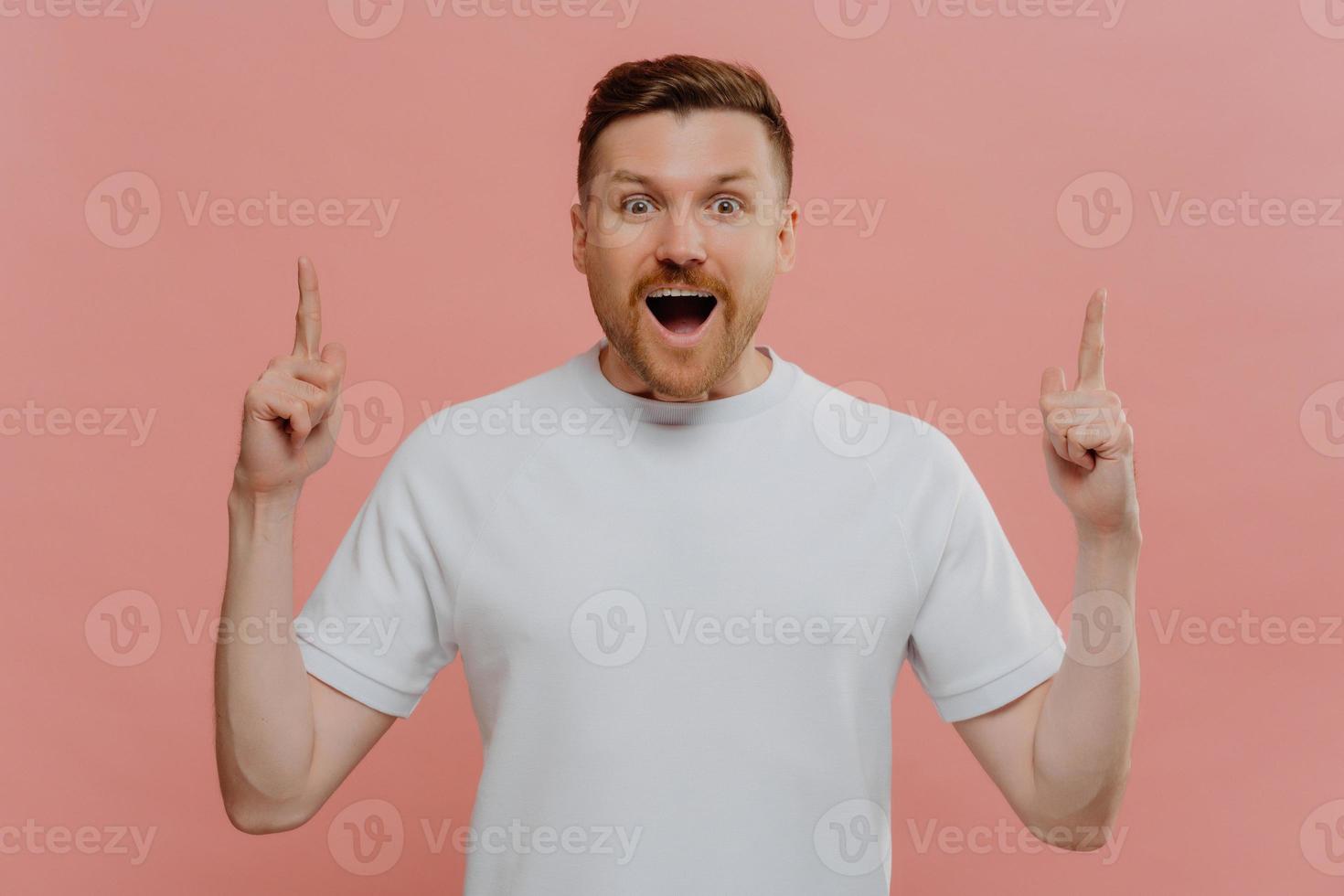 Half length shot of impressed bearded European guy points above with both index fingers gasps fromm wonder has surprised face expression wears basic white t shirt isolated over pink background photo