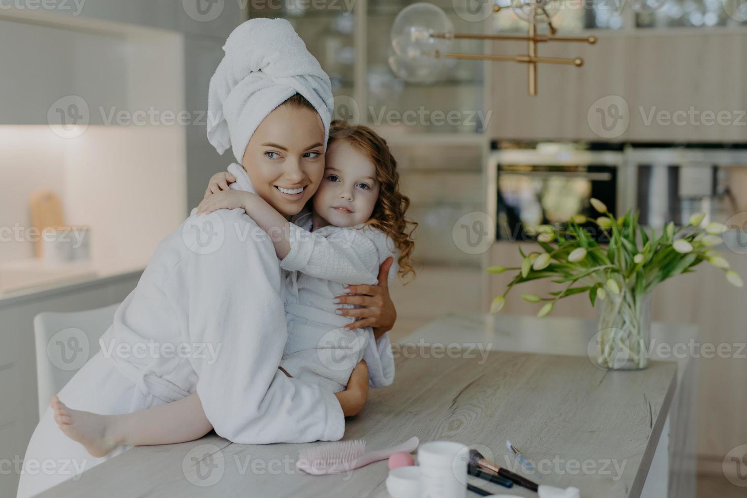 la foto de la joven madre y su hija se abrazan y se expresan amor entre sí posan en un apartamento moderno, usan suaves batas de baño blancas que van a someterse a procedimientos cosméticos y tener una piel sana. hora de la belleza