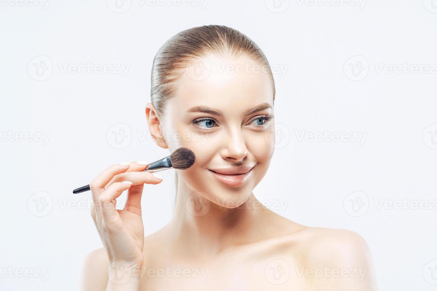 mujeres, concepto de cosmetología. tierna y saludable mujer europea da una lección de maquillaje, aplica cosméticos con un cepillo de belleza, mira a un lado con una sonrisa, aislada de fondo blanco, muestra un cuerpo desnudo perfecto foto