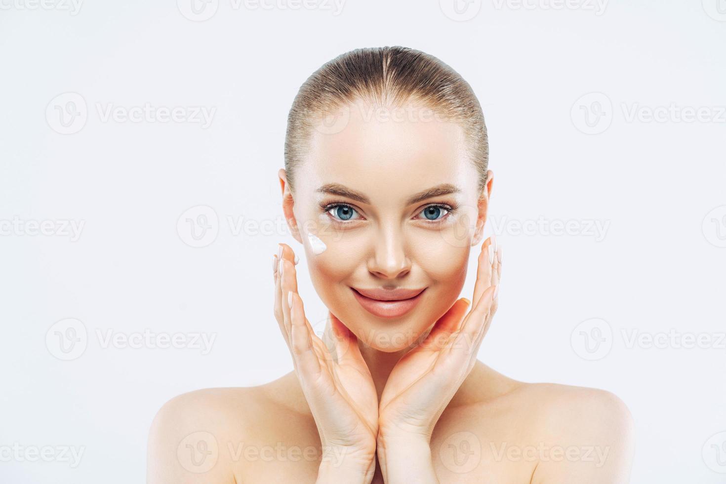 Close up shot of attractive naked young woman with combed hair, applies face cream or lotion, touches face, has natural makeup, poses against white background, looks after skin. High resolution photo