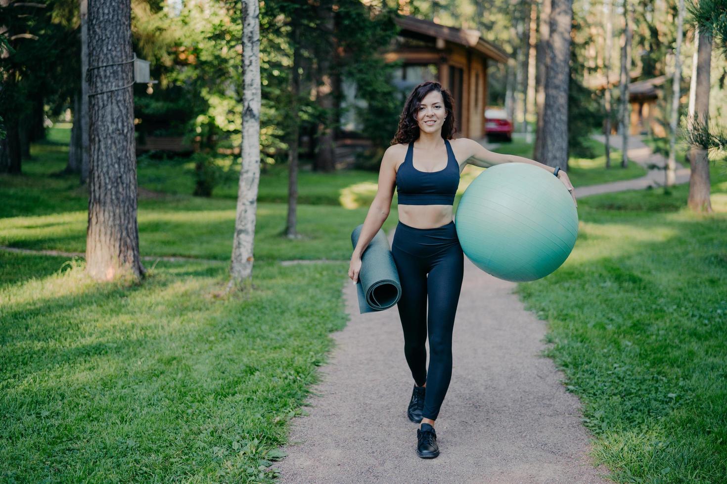 Horizontal shot of sportswoman wears cropped top and leggings, carries fitness ball and rolled up karemat, walk on road around trees and green grass, leads healthy lifestyle. Sport equipment photo