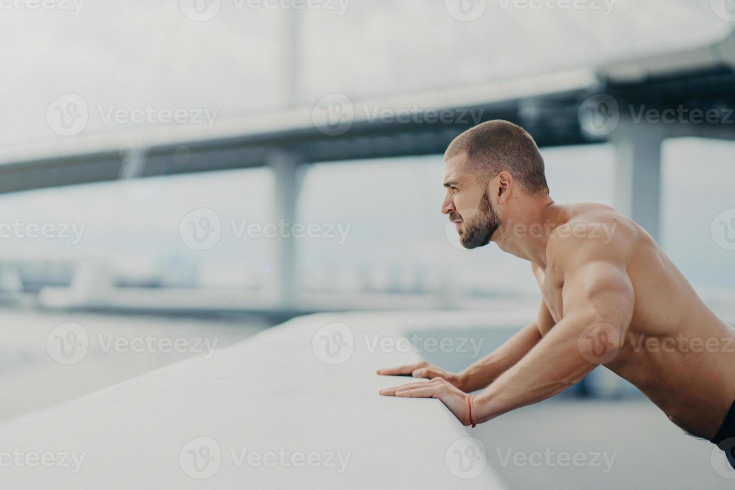 Sideways shot of handsome muscular man with thick beard does push ups exercise has workout outdoor leans on bridge fence engaged in physical activity poses with bare torso has strong muscles photo