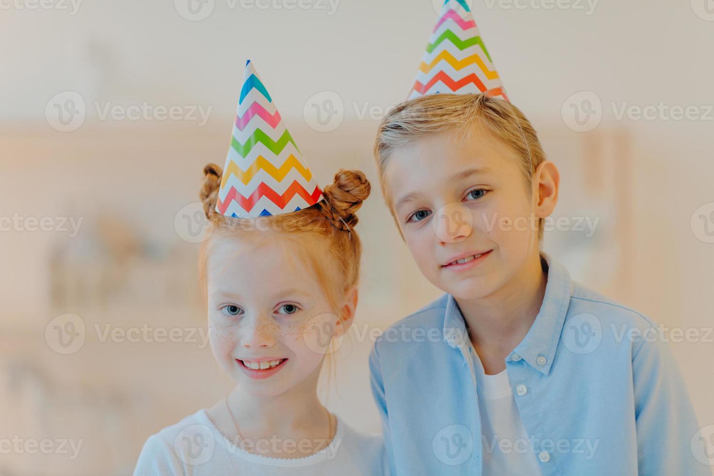 Portrait of happy ginger girl and her broghter stand closely to each other, wear party hats, come on friends birthday, look gladfully at camera, pose against blurred background. Childhood concept photo