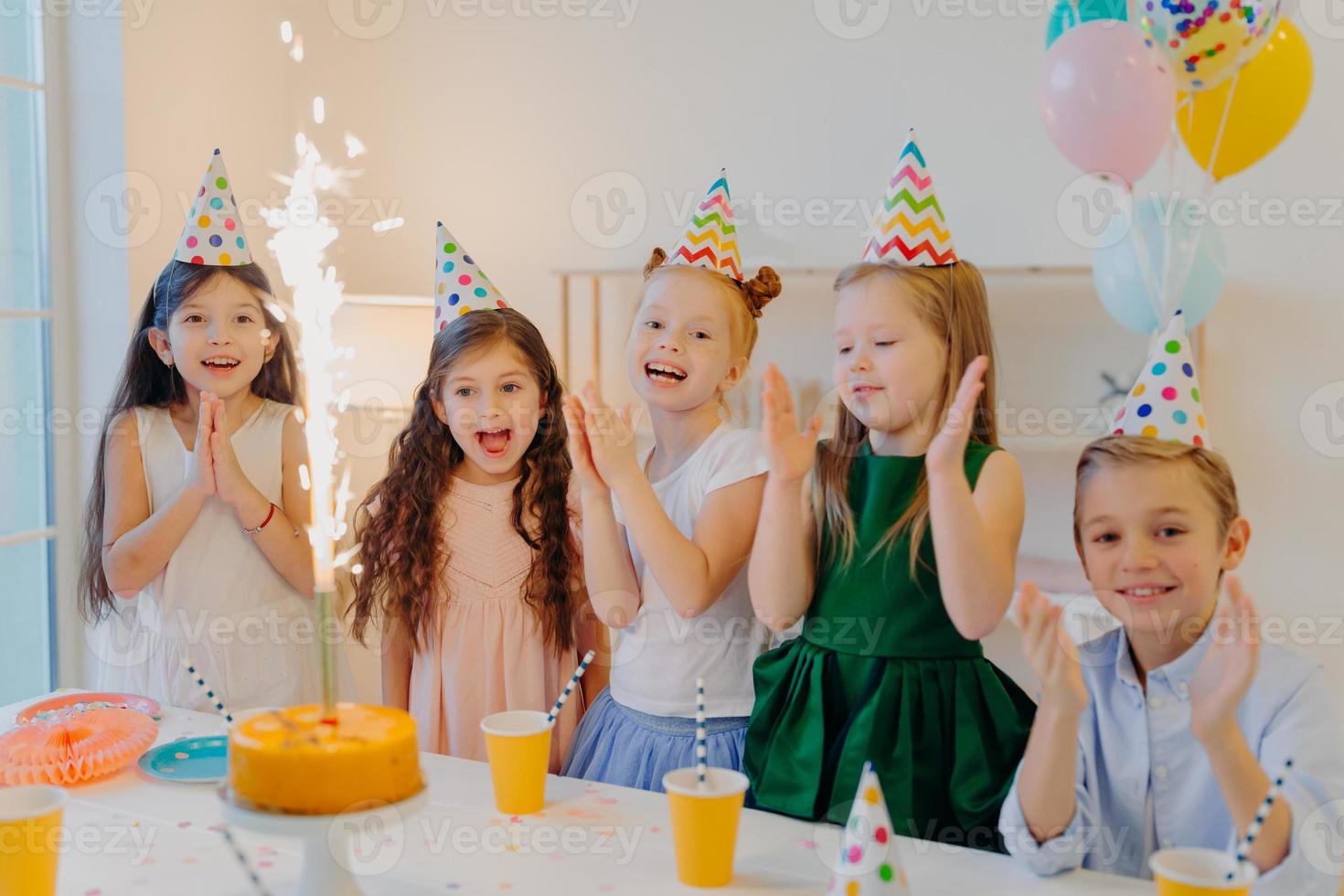 alegres amigos de cinco posan cerca de la mesa festiva con un gran pastel, una taza de bebidas, usan sombreros de fiesta, miran alegremente el brillo, ríen alegremente, posan en el interior, celebran el cumpleaños foto