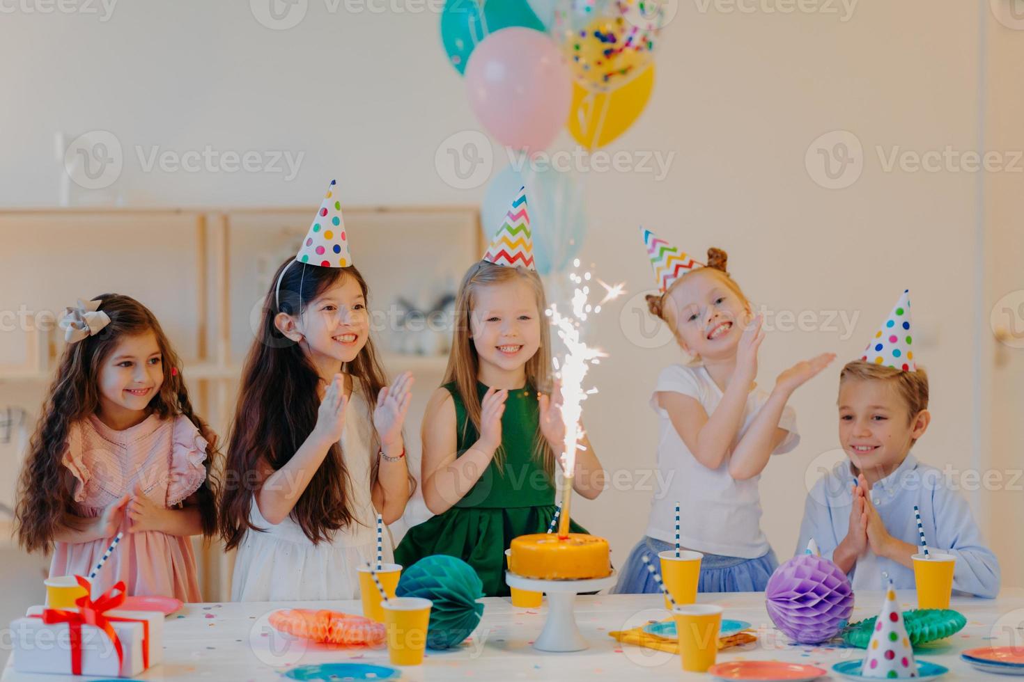 Children and holiday concept. Glad five friends look gladfully on cake with sparkle, celebrate birthday, wear party cone hats and hold air balloons, have happy expressions photo