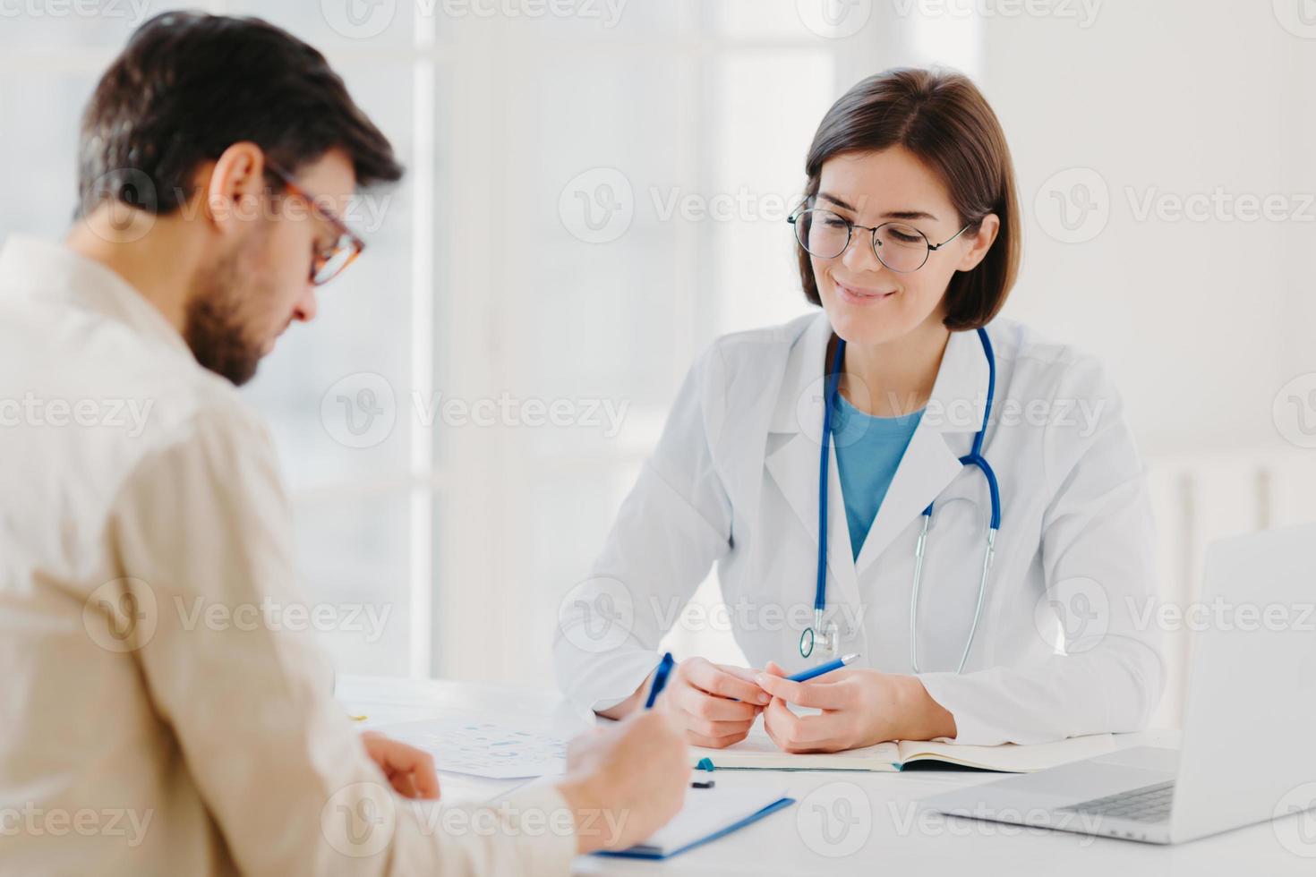 Male patient writes down application form, gets consultancy and advice about home treatment from doctor, use laptop and medical document at desk, pose at office together. Man doctors appointment photo