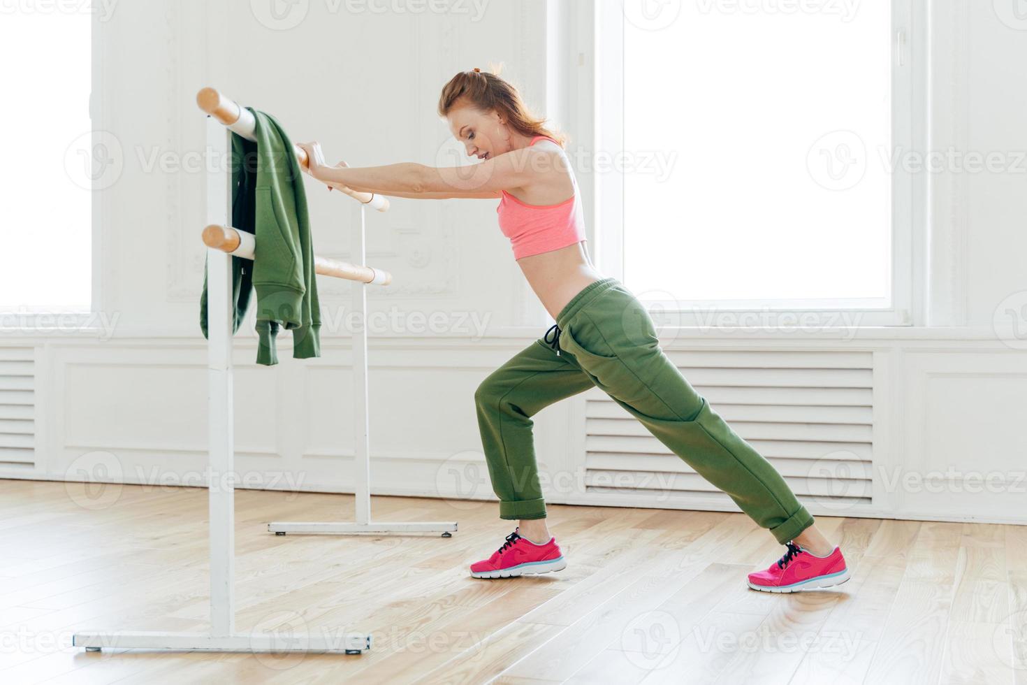 Sport, recreation, flexibility concept. Motivated strong female makes plank, leans at ballet barre, stretches before exercises prepares muscles or biceps for hard training. Aerobics instructor indoor photo