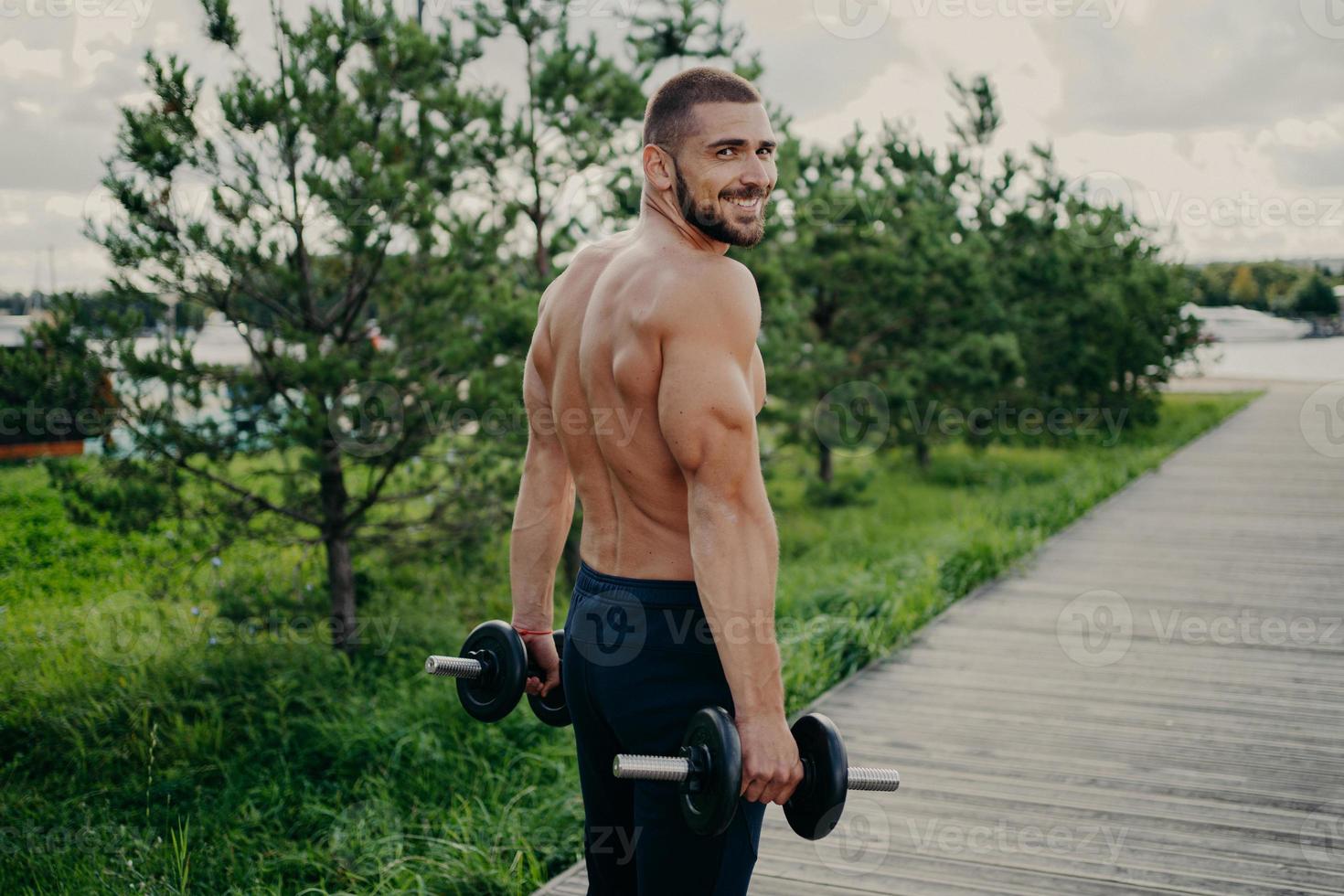 Muscular satisfied athlete bearded man gets energy during sport training, has strong arms, raises barbells, dressed in shorts, poses outdoor, enjoys regular workout in nature, builds healthy body. photo