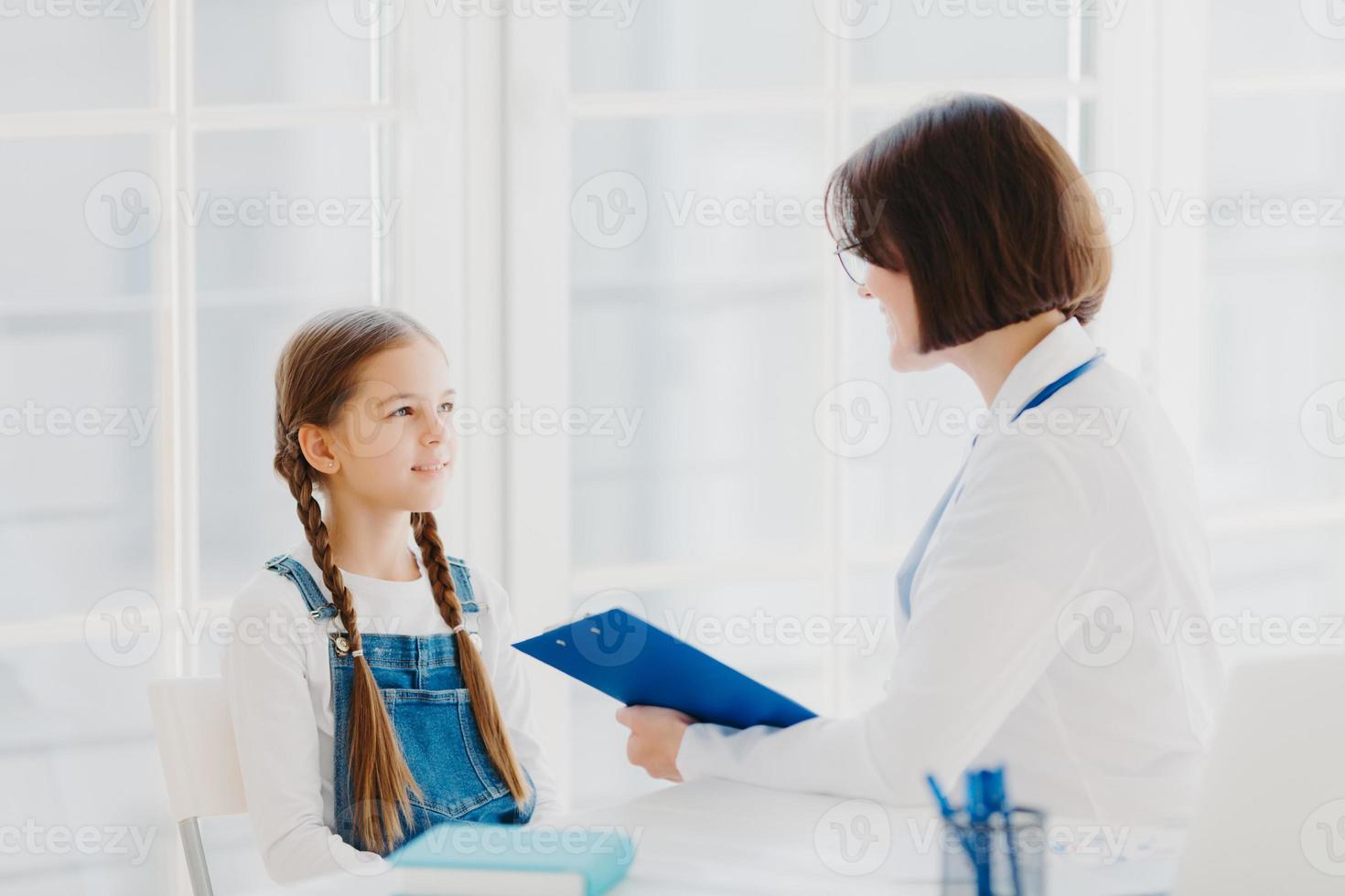 Female pediatrician examines little child, listens carefully to small kid patient, writes down notes in clipboard, pose in clinic or hospital against white window. Children healthcare concept photo