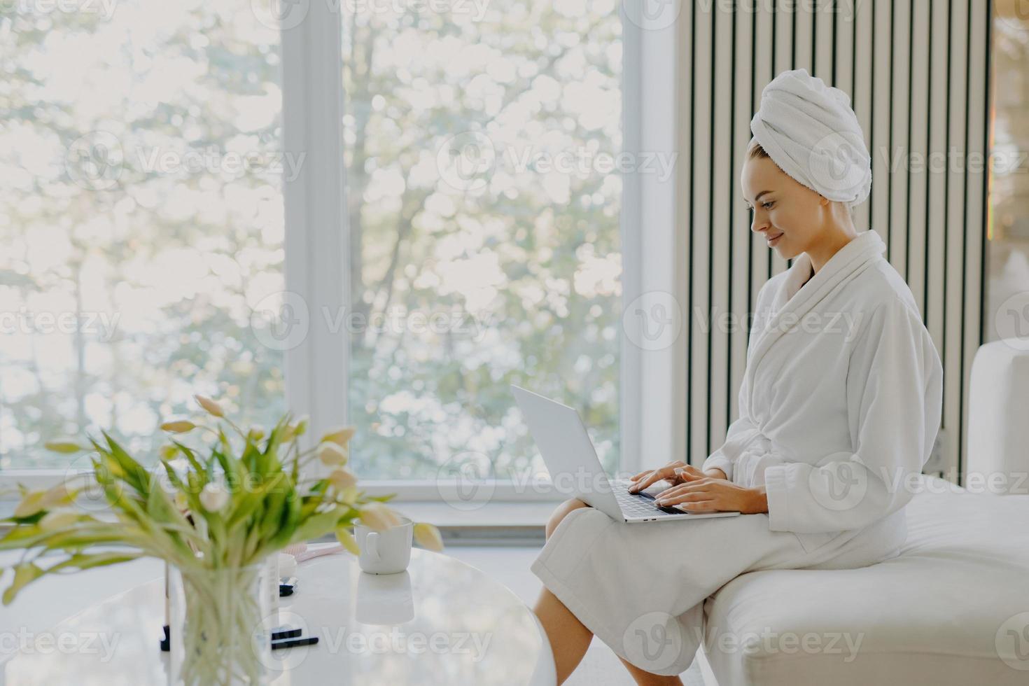 Sideways shot of attractive refreshed Caucasian woman sits at comfortbale sofa works on laptop computer wears bathrobe and wrapped towel on head poses against big window in cozy spacious room photo