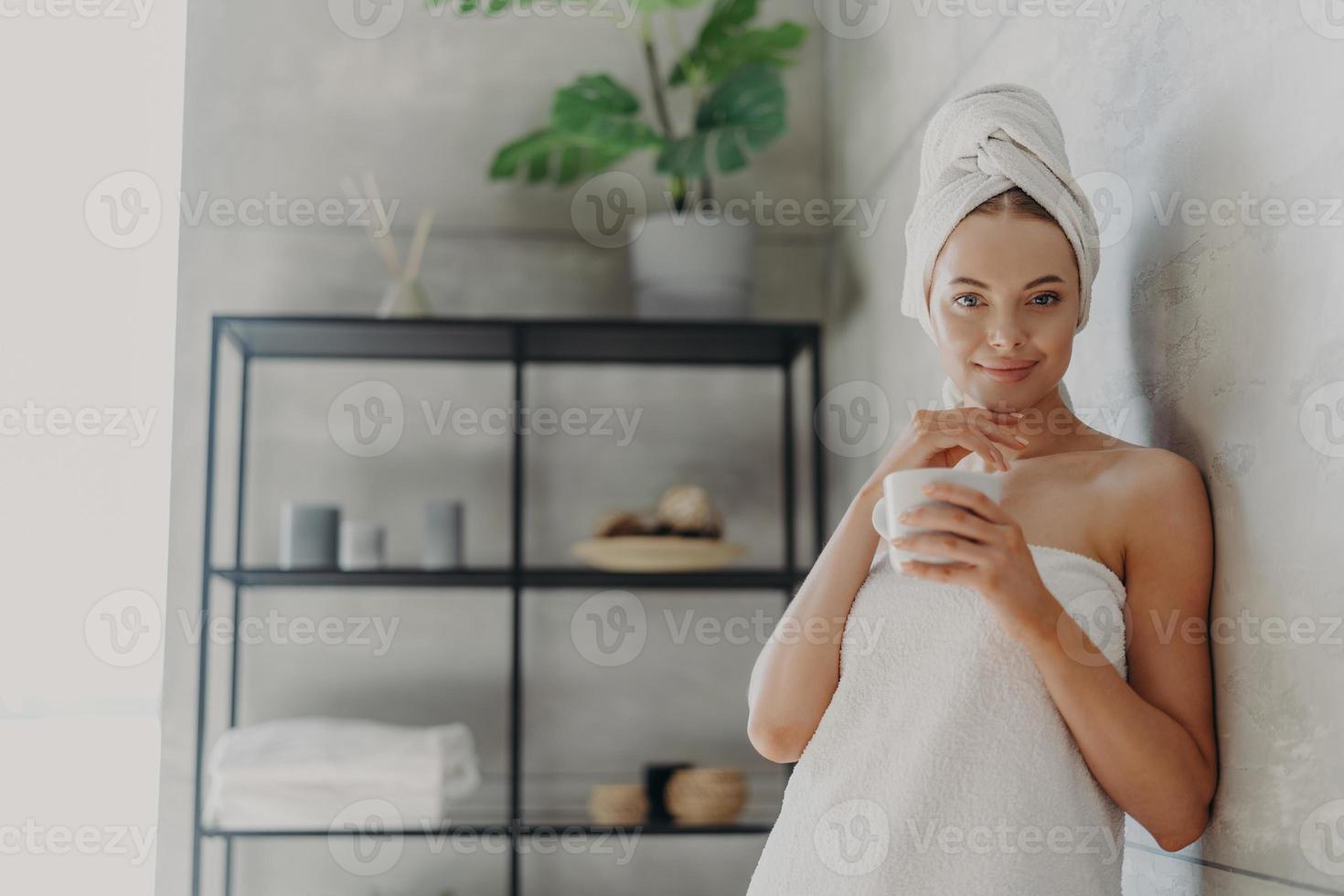 Pretty female model has perfect skin, touches jawline gently, drinks hot beverage, feels refreshed and relaxed, wrapped in bath towel, poses in bathroom. Natural beauty and skin care concept photo