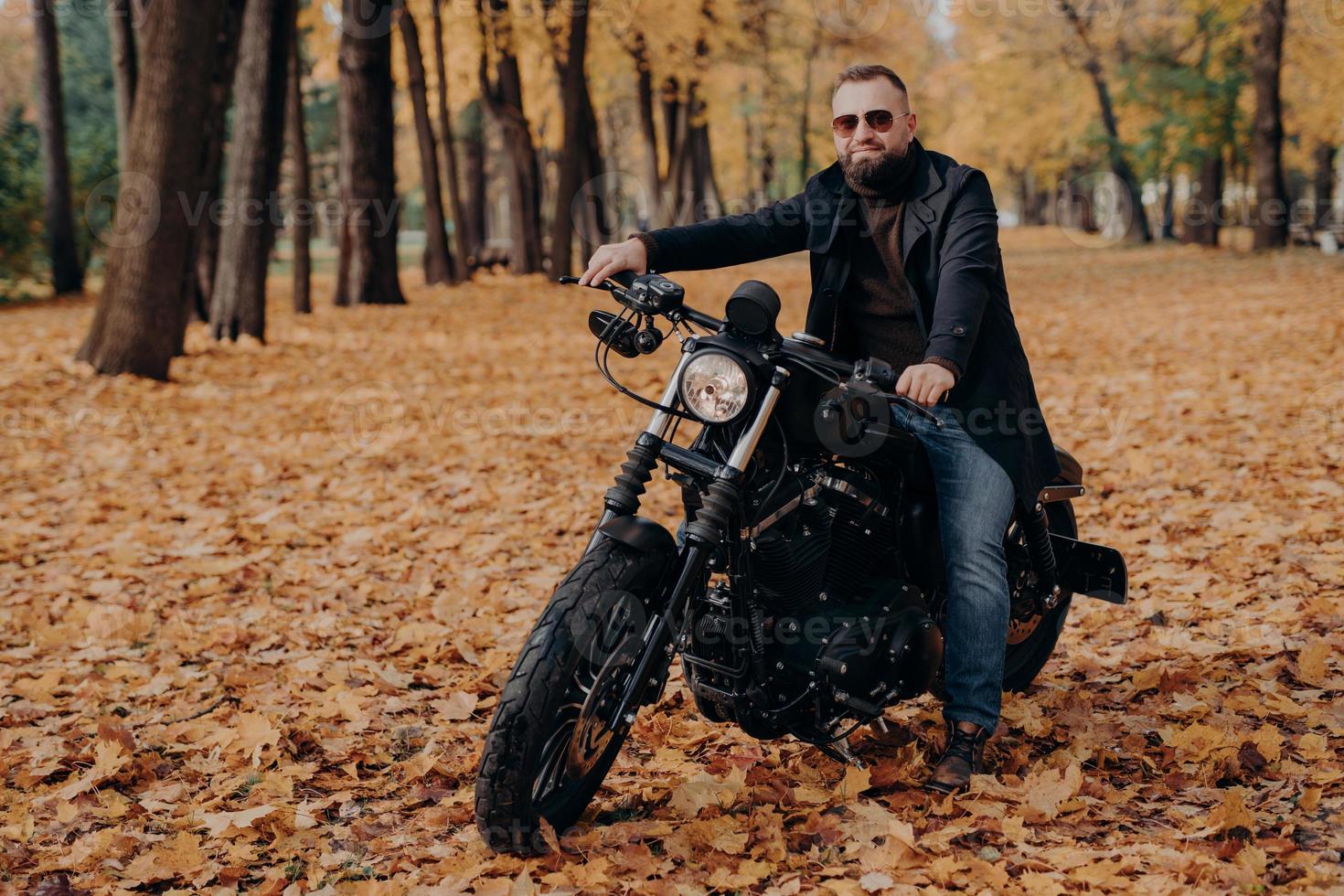 Travel, speed, freedom concept. Fashionable male motorcycle driver poses on black motorbike, wears protective sunglasses, black jacket and shoes, rides in beautiful yellow park during autumn time. photo