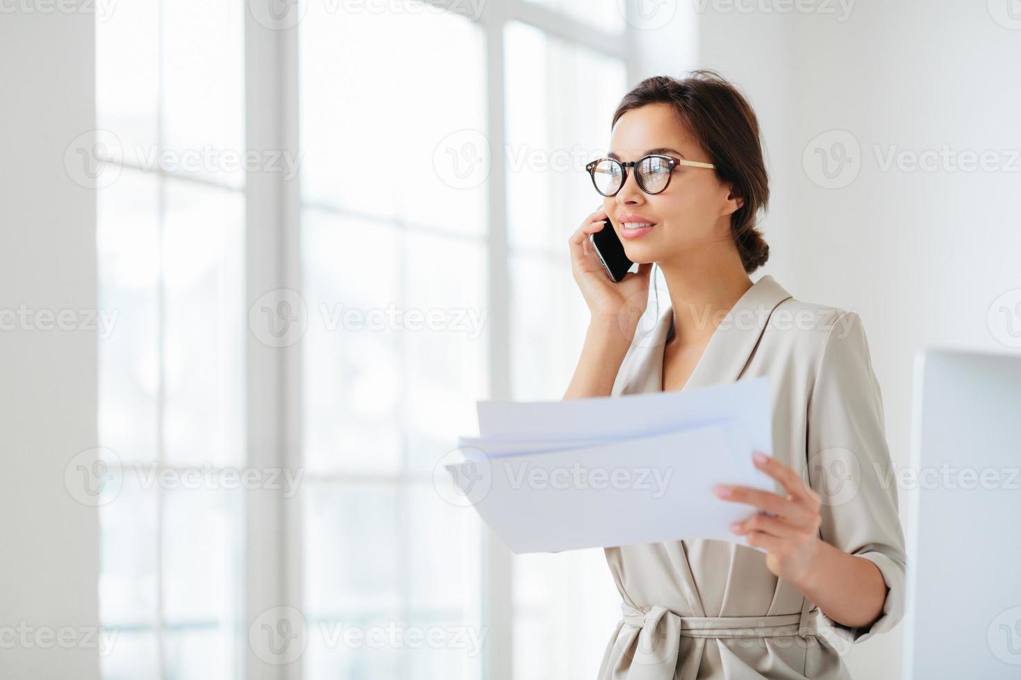 Pensive businesswoman stands sideways to camera, holds paper documents, has phone talk, prepares financial report, poses in coworking space, wears optical glasses and formal suit. Paperwork. photo