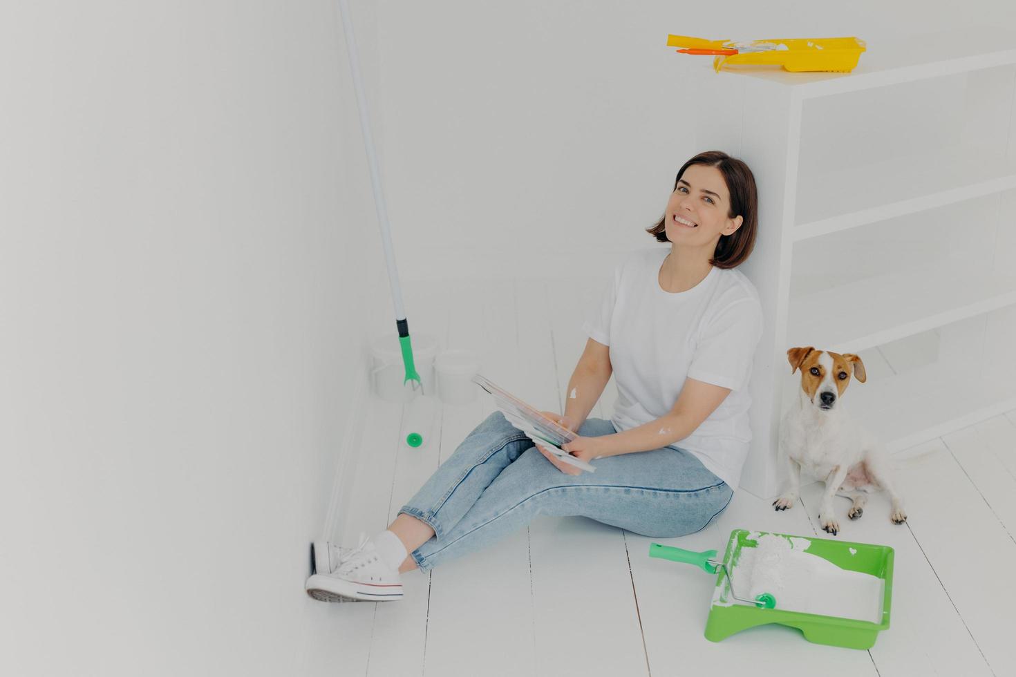 toma interior de una mujer morena feliz con ropa informal, se sienta en el suelo cerca de un cajón blanco, sostiene muestras de color, su perro jack russell terrier posa cerca, ocupado con la renovación de paredes en una casa comprada foto