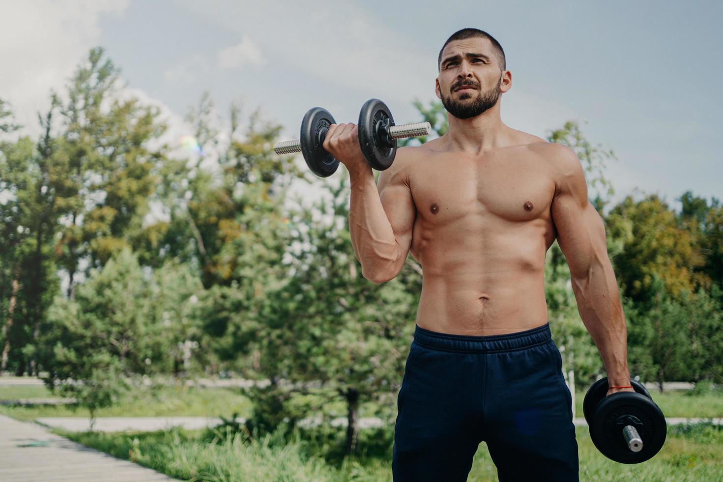 Horizontal shot of topless fitness unshaven European man has muscular body, raises barbells, wears shorts, demonstrates strong arms, has workout outdoor in park. Sportsman makes weightlifting photo