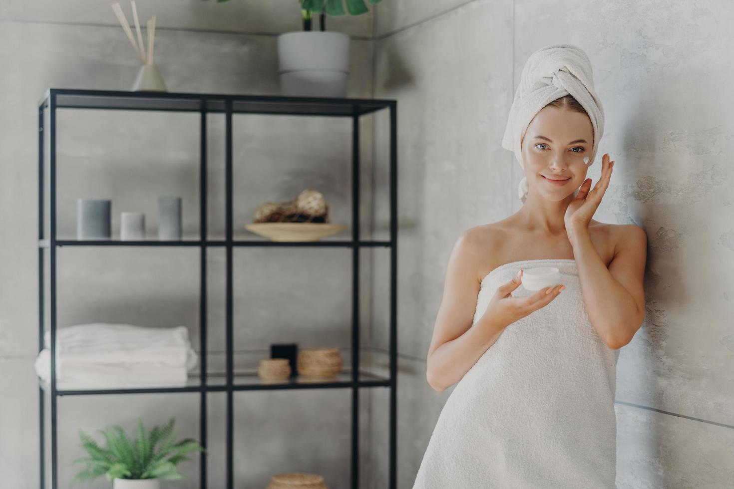 la toma horizontal de una joven modelo femenina saludable usa crema hidratante, sostiene un frasco de producto cosmético, envuelto en una toalla después de la ducha, se para cerca de la pared en un baño acogedor. procedimientos de cuidado de la piel foto