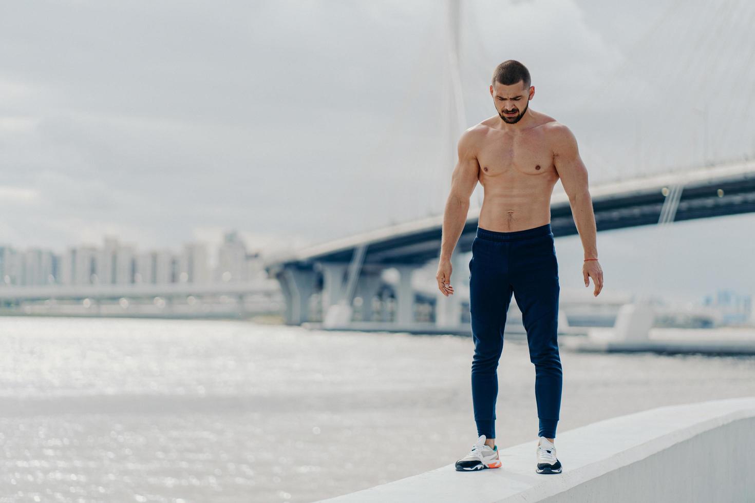 Sporty bearded man with muscular body walks near river, dressed in sport trousers and sneakers, looks down, has muscular body, stands against beautiful landscape. Athlete bodybuilder poses outside photo