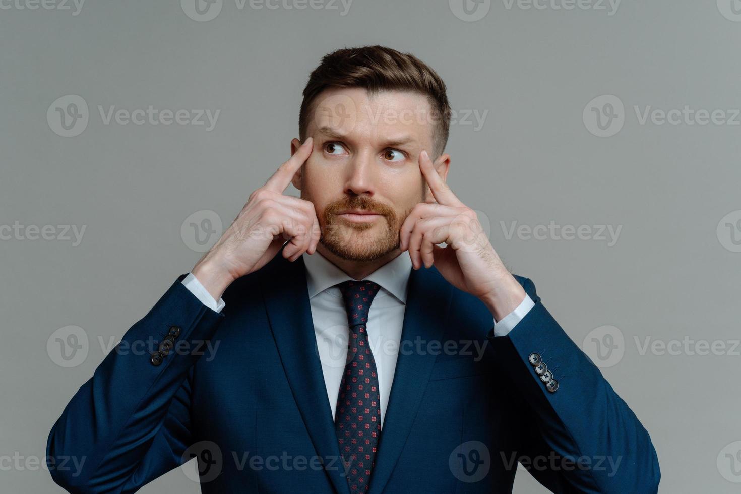 Indoor shot of motivated thoughtful bearded businessman keeps fingers on temples looks away gets ready for conference dressed in formal clothes isolated over grey background has hard working day photo