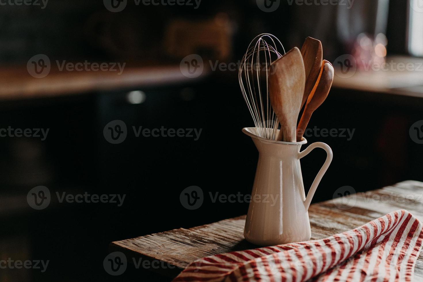 Kitchen accessories on wooden table. Utensils in white ceramic jar against dark background. Rustic style. Dishware for preparing meal. Wooden spoons shpatula and whisk photo