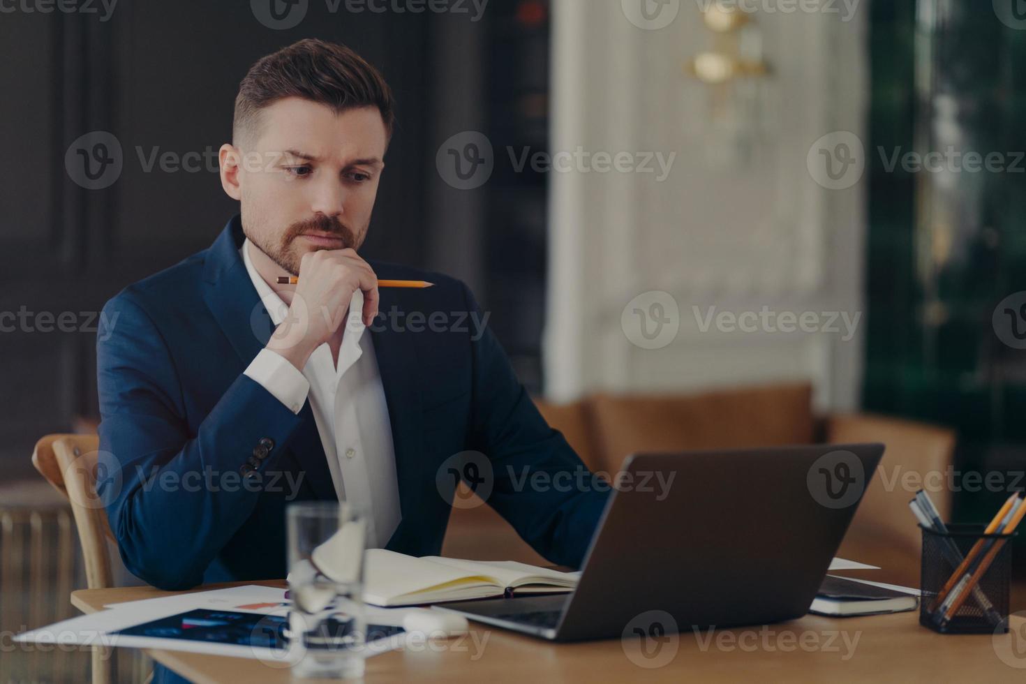 Serious male entrepreneur holds chin makes notes in diary works on laptop computer makes money online dressed formally writes information. Confident managing director involved in working process photo