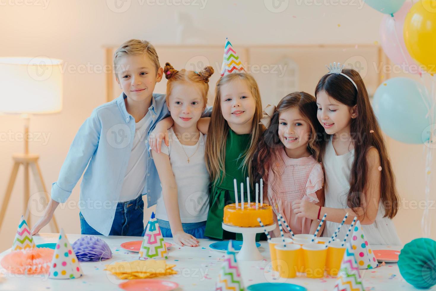 Friendly children embrace while pose near festive table, blow candles on cake, have party mood, celebrate birthday or special occasion, have joyful expressions. Childhood, fun and entertainment photo