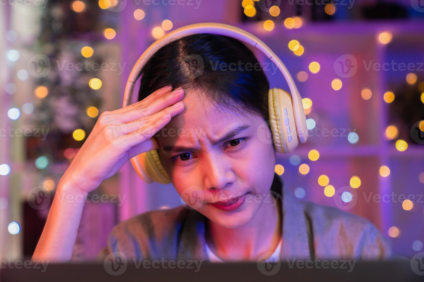 Stressful young Asian woman wearing headset and sitting on the chair with a pile of papers document on the table and looking on the computer screen on night. photo