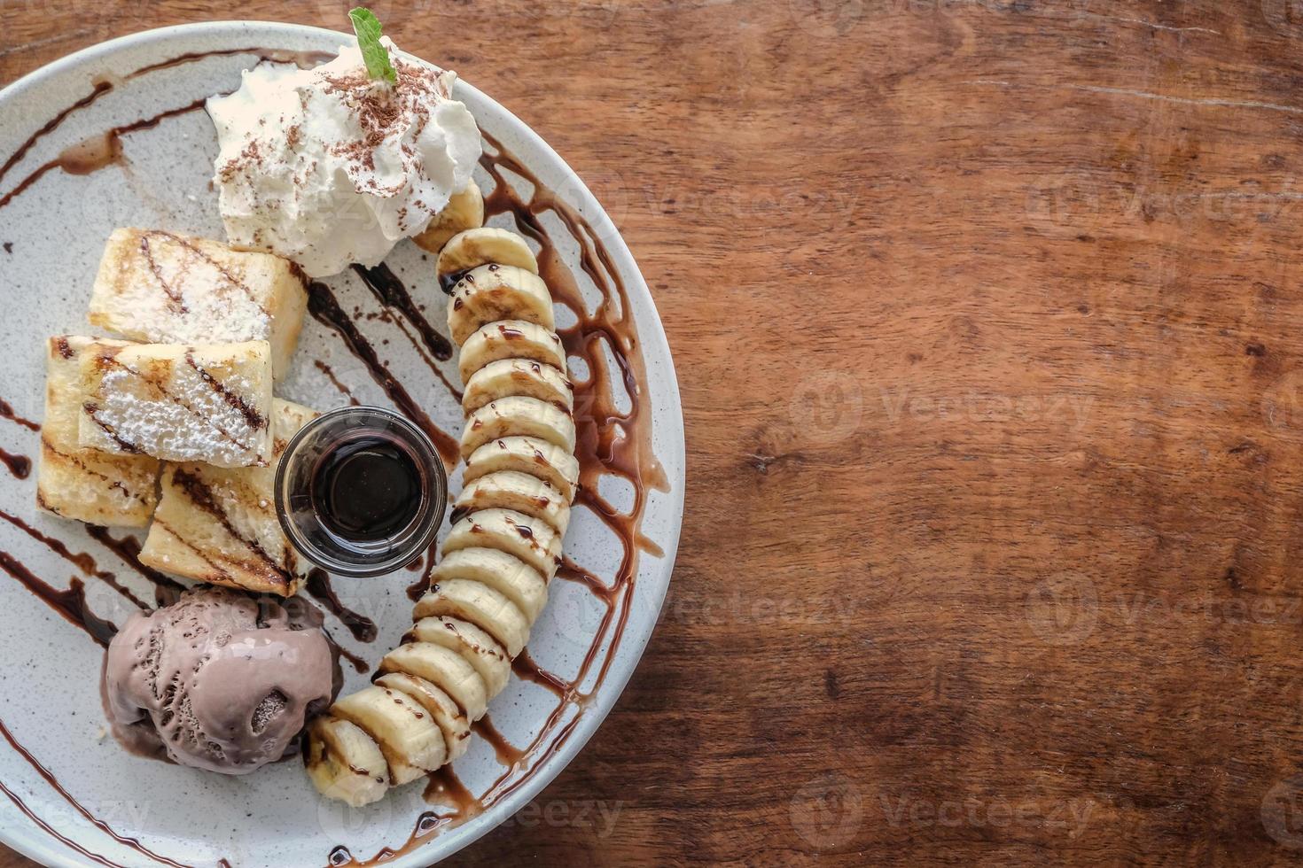 Top view of toast, chocolate ice-cream, banana slices and whip cream in white plate, all dripped with chocolate sauce put on wood table background. photo