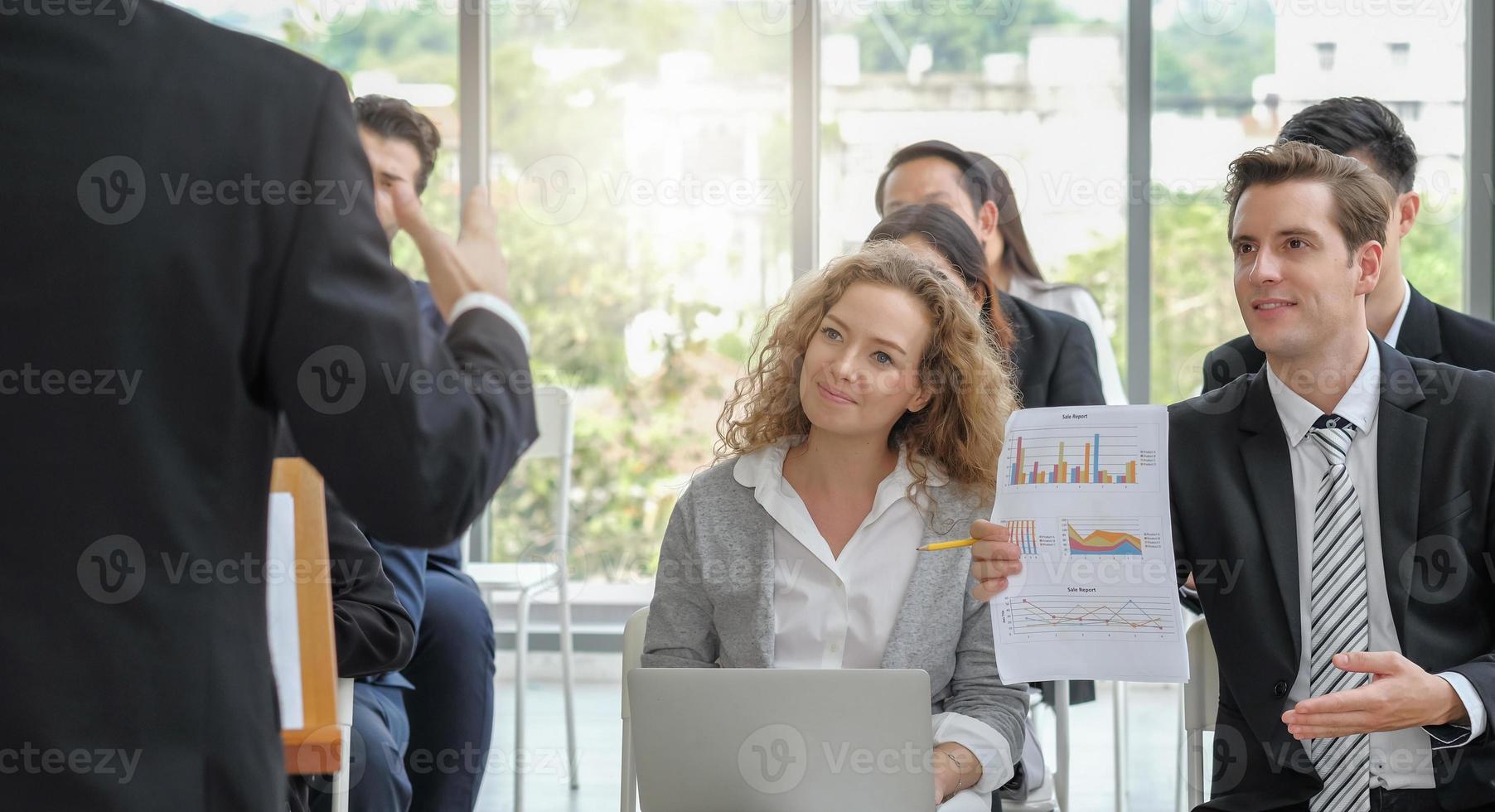 Young adult caucasian business man asking question with report in hand to speaker while making speech among the audience in corporate seminar event. Light effect. photo