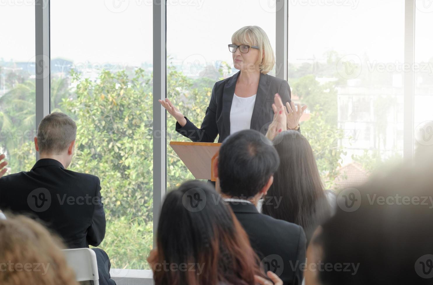 Senior business woman received applause from the audience while making speech in corporate seminar event with light effect. Selective focus. photo