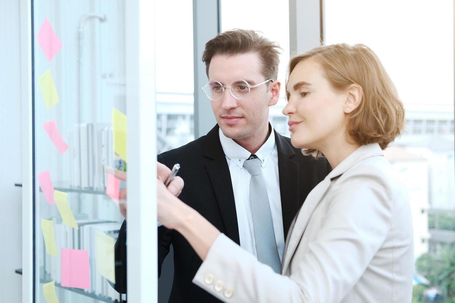 Successful businessman and businesswoman writing good ideas on sticky notes pasted on glass wall in modern office meeting. photo