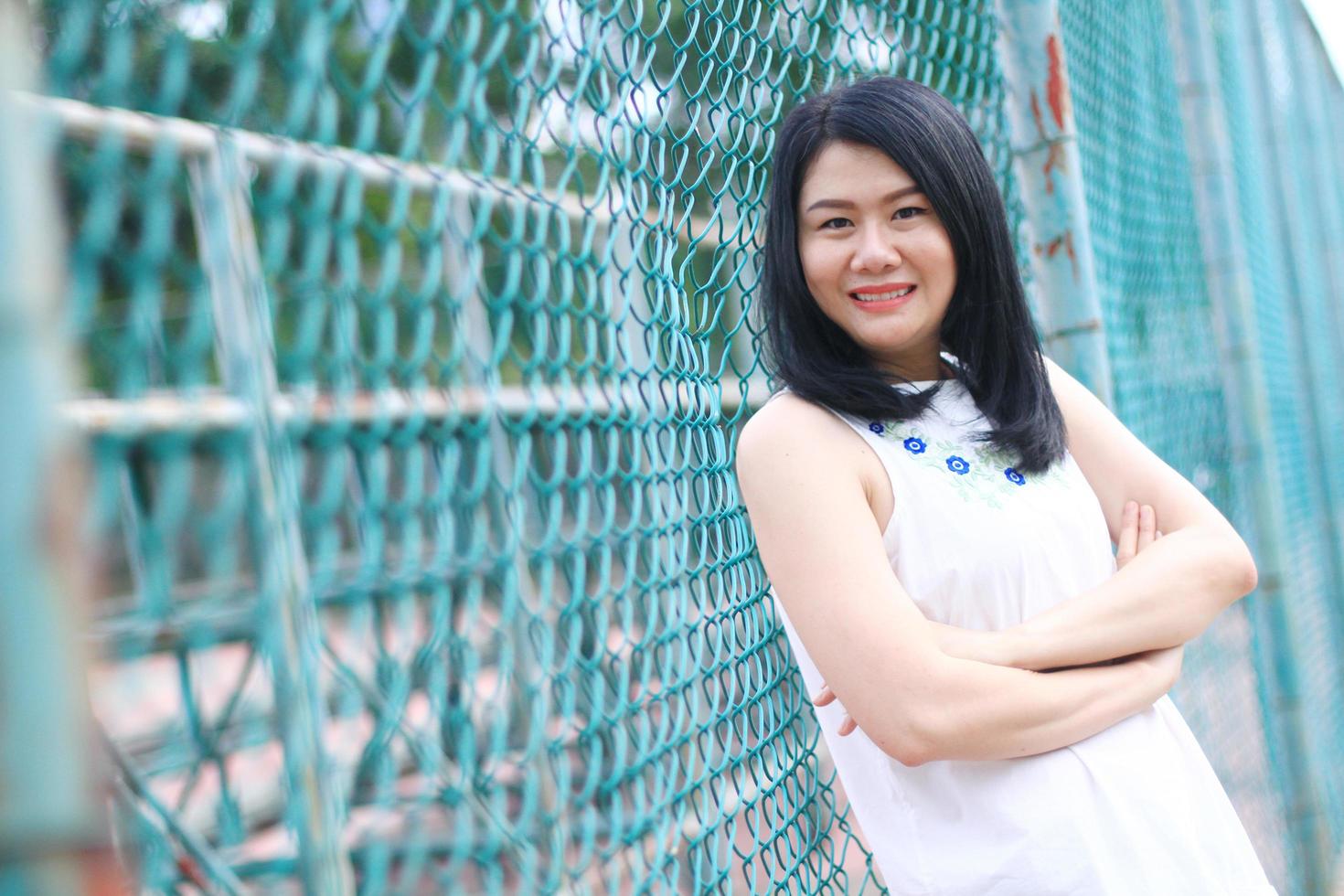 hermosa mujer asiática con vestido blanco sonriendo y de pie con una valla de acero azul en el parque natural. joven tailandesa disfruta de vacaciones con la luz del sol en el jardín. foto