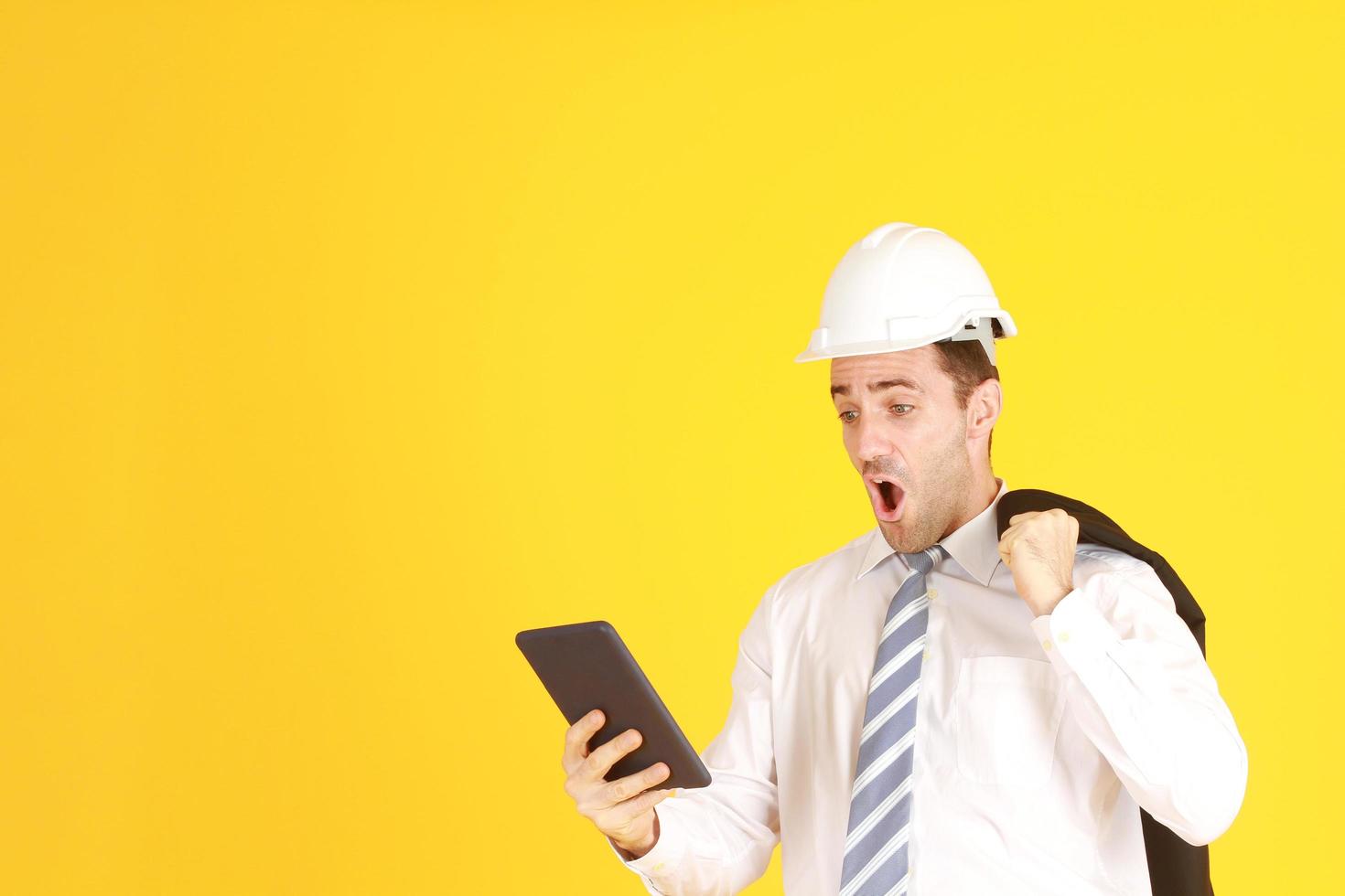 ingeniero guapo e inteligente con traje y camisa blanca y con un sombrero blanco de ingeniería de seguridad con la mano sosteniendo un teléfono inteligente sorprendido y estresado con el trabajo aislado en el fondo amarillo. copie el espacio foto