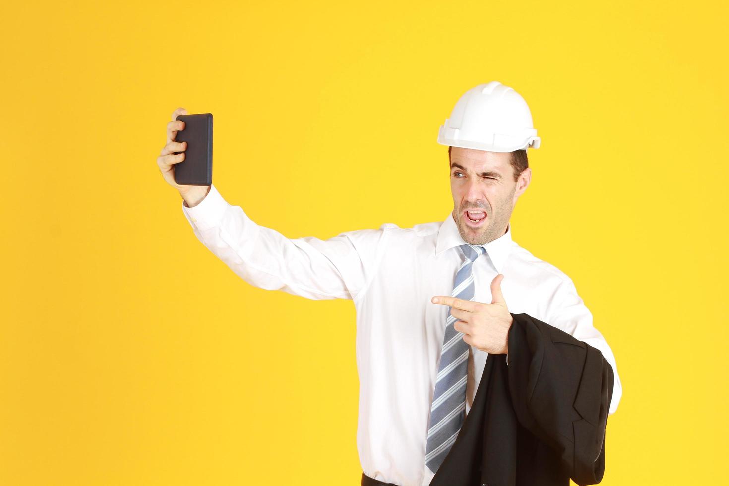 Handsome smart engineer or architecture is Wearing a white safety hat with hand holding smartphone and enjoy with Selfie by himself isolated on yellow background. Copy Space photo