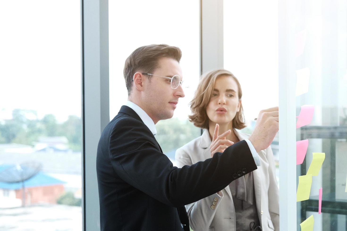 Successful businessman and businesswoman writing good ideas on sticky notes pasted on glass wall  in modern office meeting. photo