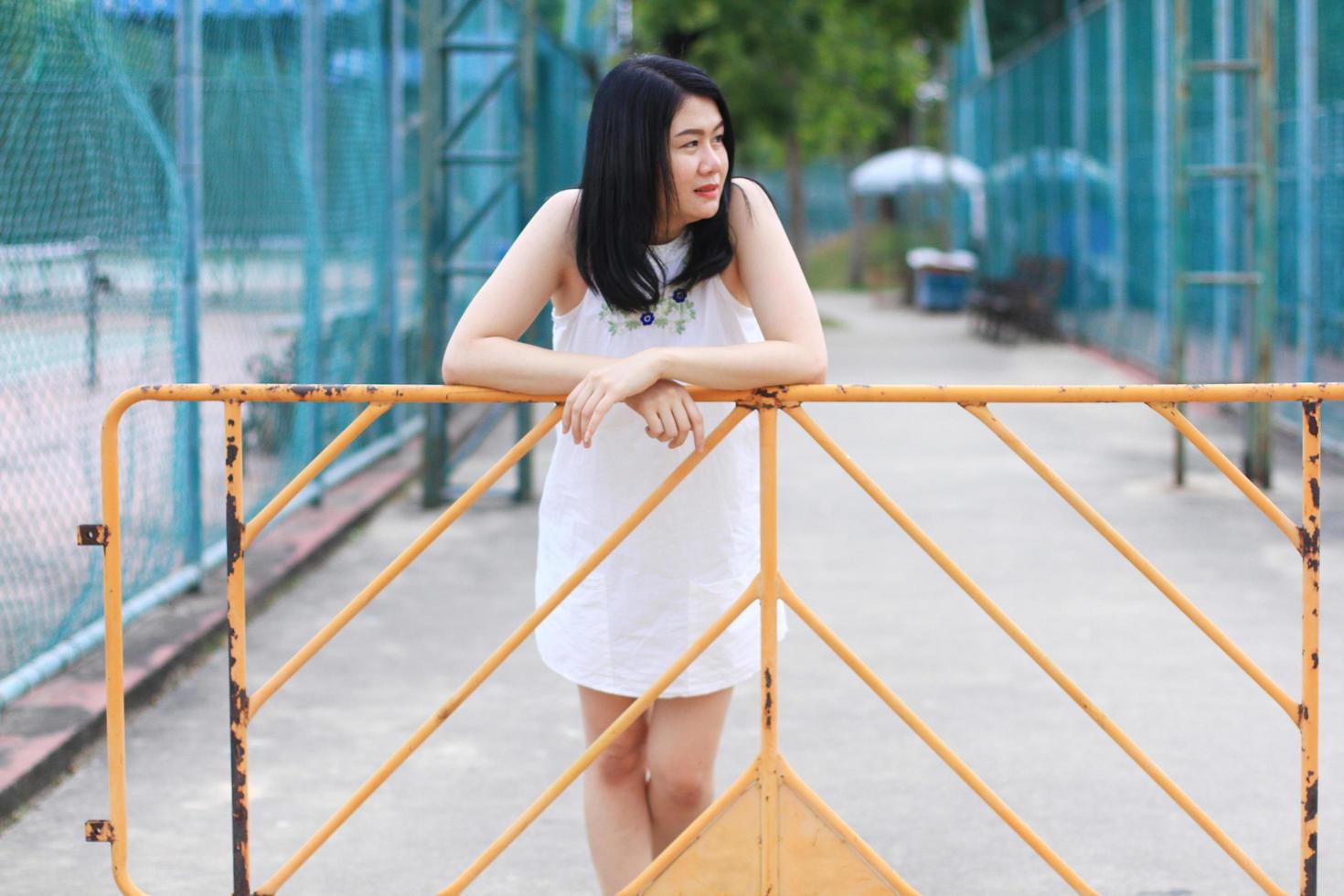 Beautiful asian woman in white dress smiling and stand with yellow steel barrior and blue grating iron fence in natural park. Young Thai girl enjoy on holiday with sunlight in the garden. photo