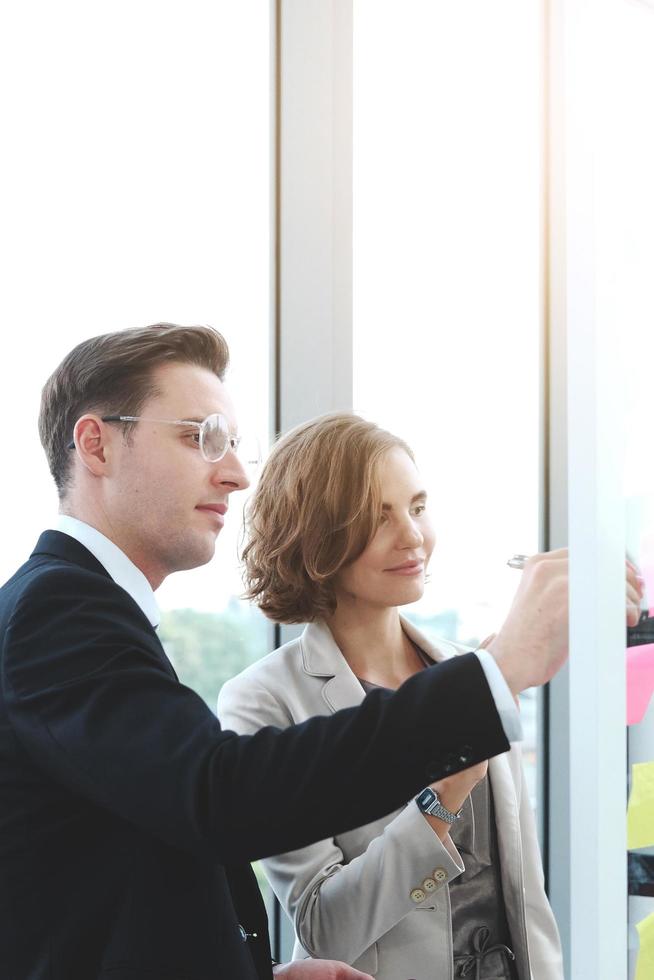 Successful businessman and businesswoman writing good ideas on sticky notes pasted on glass wall  in modern office meeting. photo