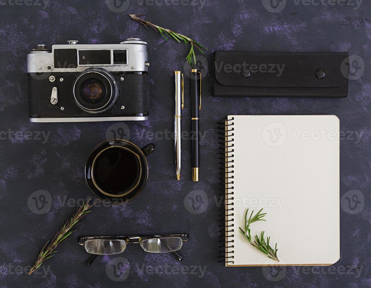 Flat lay, top view office table desk. Desk workspace with retro camera, diary, pen, glasses, case, cup of coffee, rosemary on dark background. photo