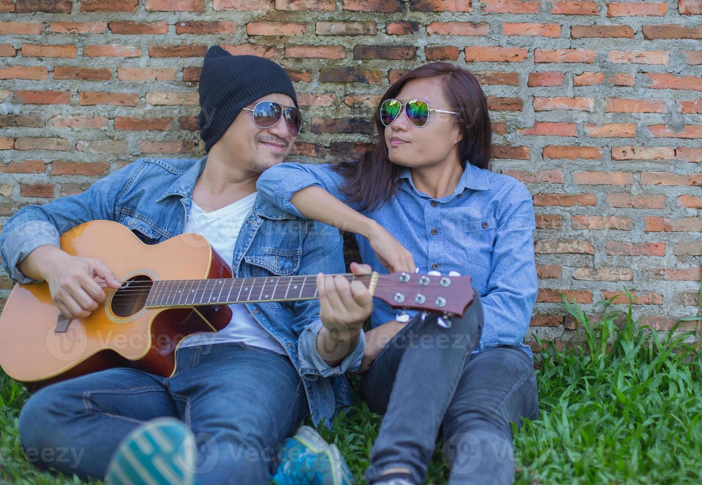 Hipster man playing guitar for his girlfriend outdoor against brick wall, enjoying together. photo