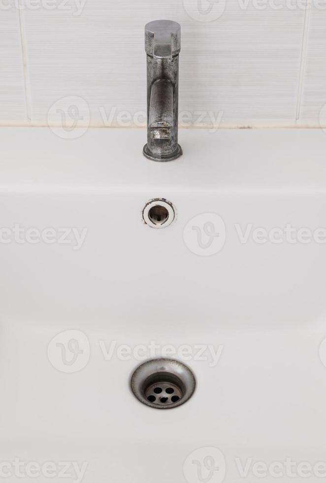 The ceramic sink basin with the dirty faucet near the white wall of the restroom. photo