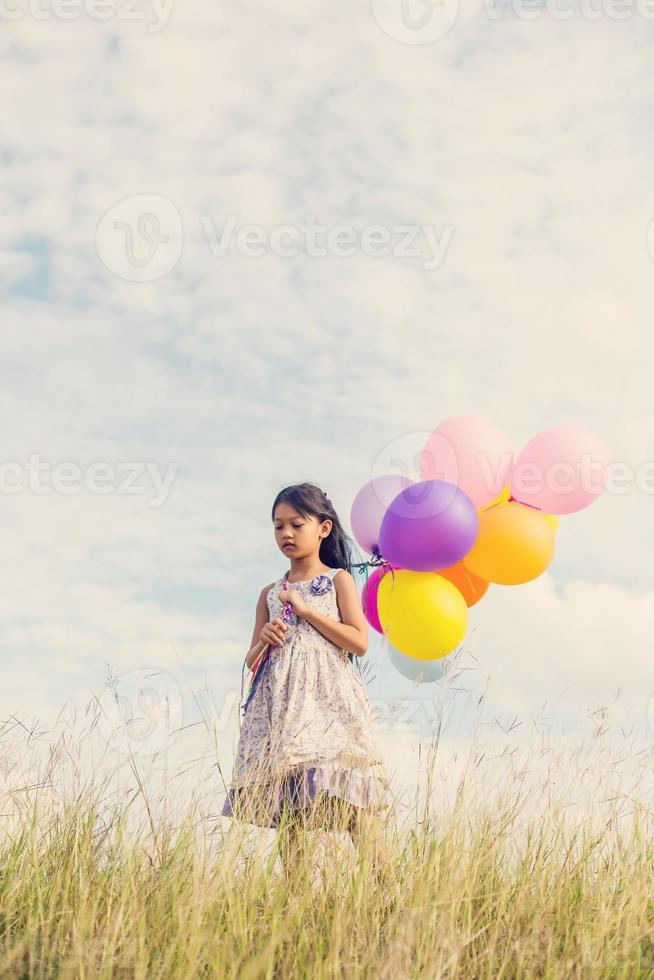 Cute little girl holding colorful balloons in the meadow against blue sky and clouds,spreading hands. photo